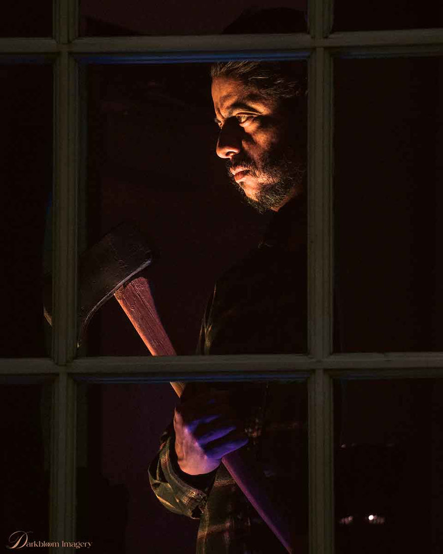 Man holding foam axe prop behind window with dramatic dark lighting.