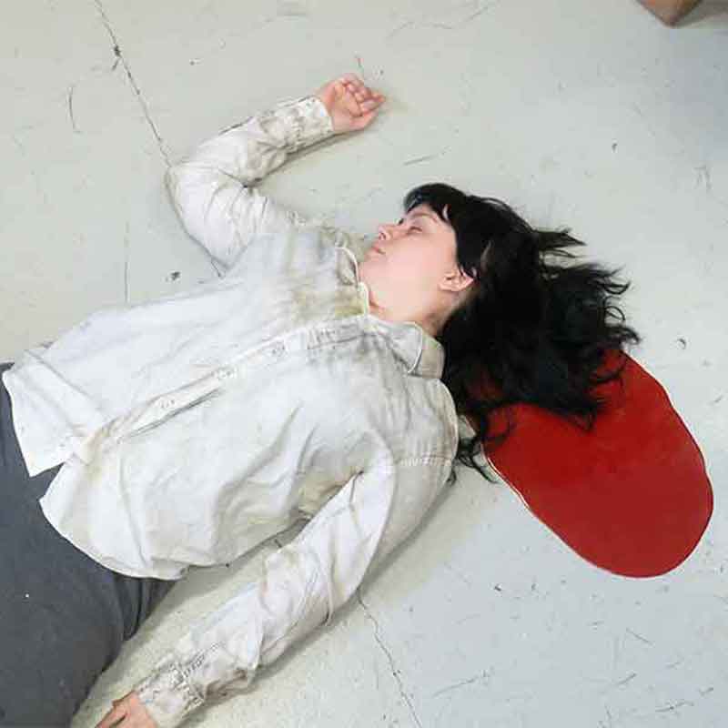 Woman in a dirty white button-up shirt lying in a puddle of fake silicone blood on the floor.